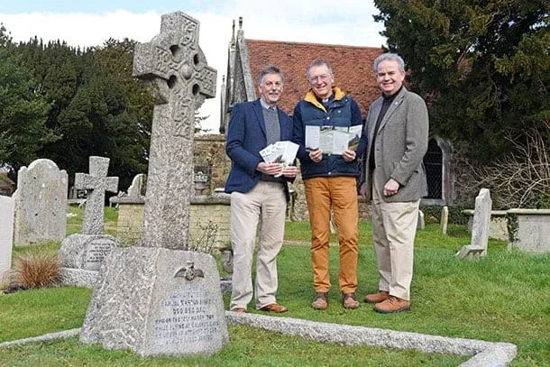 Three men stand in a graveyard beside a large Celtic-style gravestone, holding leaflets. Tombstones and a church with a red roof are visible in the background. The grave is dedicated to a First World War flying ace. The weather appears clear and mild.