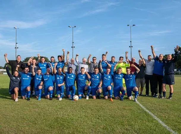 A football club in blue kits poses on a grassy pitch with coaches and staff, all raising their arms and smiling for a group photo under a clear blue sky, grateful for the recent donation that helped them see the light.