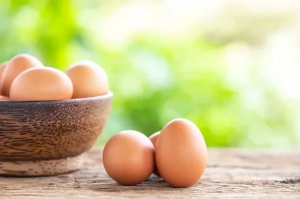 A brown wooden bowl filled with eggs of varying egg size sits on a rustic wooden surface outdoors, with three eggs beside the bowl and a blurred green background.