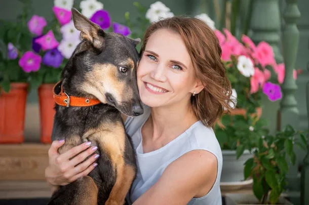 A smiling woman with light brown hair cuddles her black and tan dog wearing an orange collar. Surrounded by colourful flowers in pots, these happy dog owners enjoy a cheerful and vibrant background.