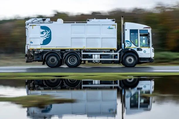 A white rubbish lorry with a deer logo on the side drives quickly past a reflective body of water, showing its mirrored image. Trees with autumn foliage are blurred in the background.