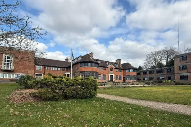 A large brick building with multiple wings, surrounded by grass, shrubs, and trees under a partly cloudy sky. There are tables and benches on a patio area in front of the building.