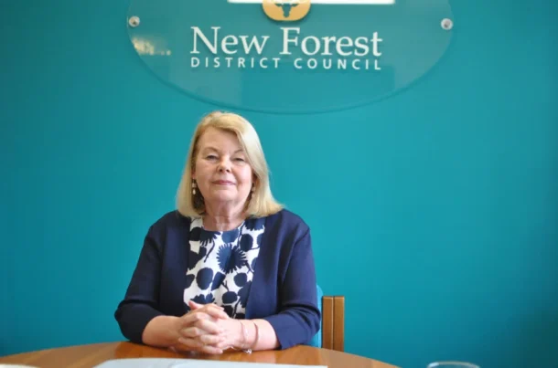 A woman with blonde hair sits at a table, hands clasped, in front of a teal wall with a New Forest District Council sign. She wears a navy blazer over a patterned top and looks at the camera.