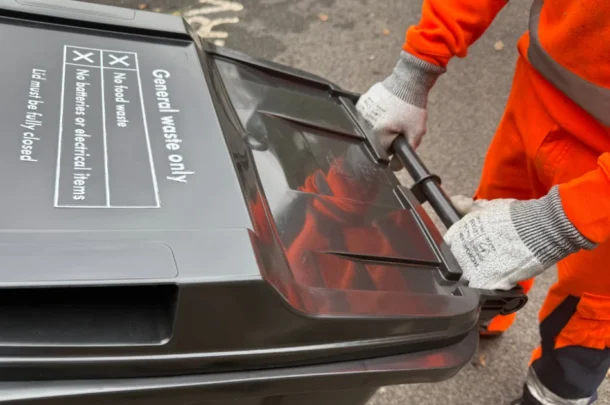 A person wearing orange work clothes and grey gloves is wheeling a black general waste bin labelled for no food, batteries, or electrical items on a paved surface.