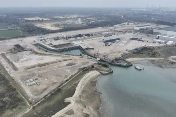 Aerial view of an industrial waterfront area with docks, construction materials, and buildings near the shoreline. Sparse vegetation and several access roads are visible, with water leading to a small harbour.