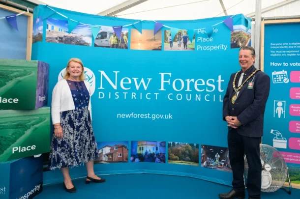 Two people stand smiling in front of a New Forest District Council display, featuring banners, photos of local scenes, and large cubes labelled People, Place, and Prosperity.