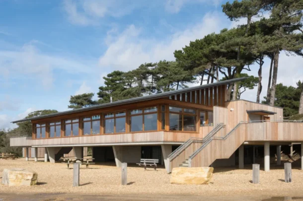 A modern wooden building with large windows, elevated on stilts, sits among trees in Lepe Country Park. There are outdoor stairs, picnic tables underneath, and a gravel area in the foreground under a partly cloudy sky.