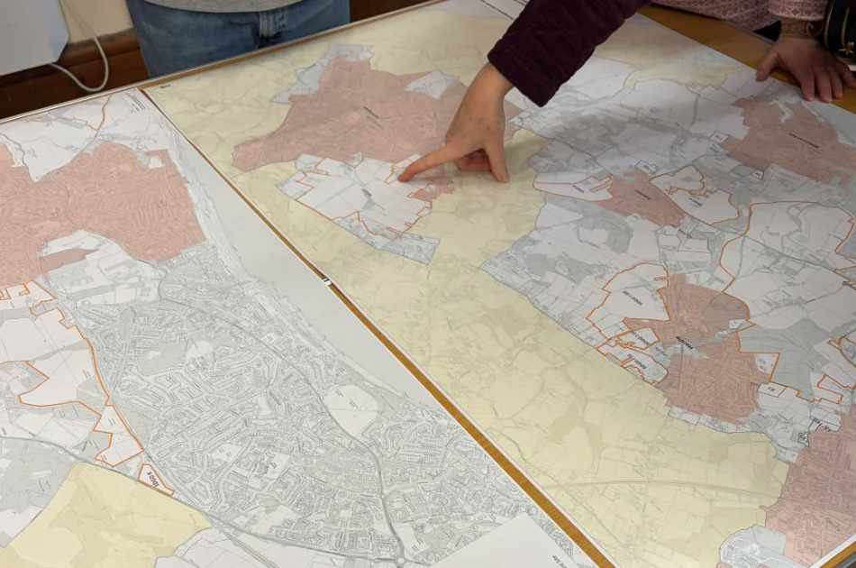 A person points at a specific area on a large map spread out on a table, with another person standing nearby. The map shows different regions shaded in various colours and outlined in orange.