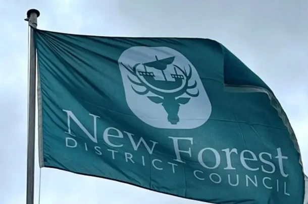 The teal flag of the New Forest District Council, featuring a white emblem of a deer’s head topped with a ship, waving on a flagpole.